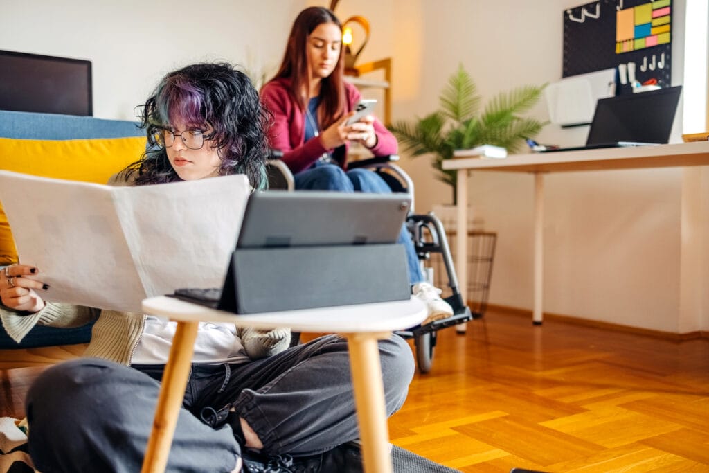 Young female adult with disability preparing for exam studying together with friend at home who is looking at charts while sitting on floor in front of digital tablet looking at paperwork from complex home modificaiton builder