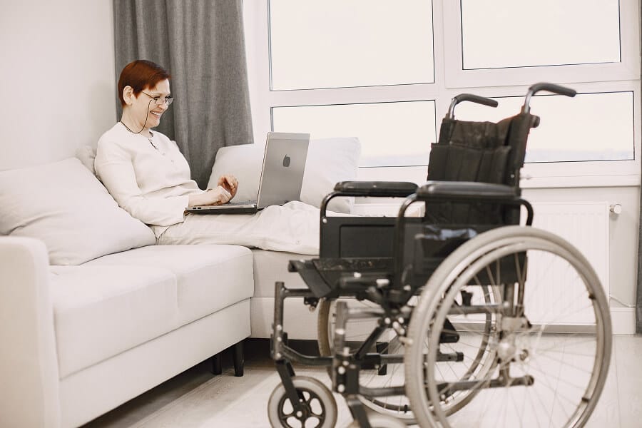 A woman sitting on the sofa using her laptop. A wheelchair is nearby.